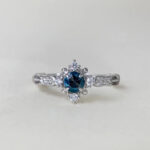 Swan Pırlanta London Blue Topaz tektaş yüzük merkezinde derin mavi topaz taşı, çevresinde beyaz pırlantalar ve ince zarif kesimlerle çevrelenen gümüş tonlu yüzük tasarımı